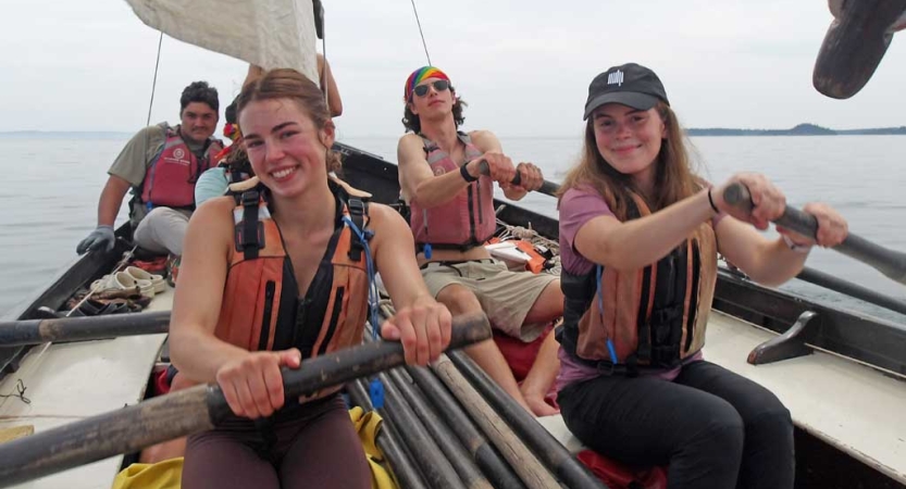 Four people wearing life jackets sit in a boat. Three of them are using oars. 